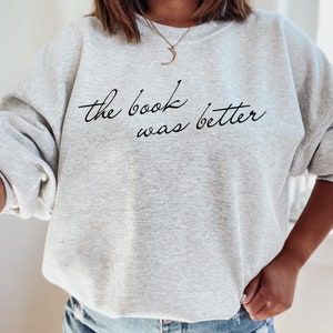 Puede incluir: Una sudadera gris claro de cuello redondo con el texto negro en cursiva "the book was better" impreso en el pecho. La prenda tiene mangas largas y un cuello acanalado, ofreciendo un aspecto informal con un mensaje claro y legible.