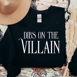 K&ouml;nnte beinhalten: Schwarzes Sweatshirt mit dem wei&szlig;en Text "DIBS ON THE VILLAIN". Das Sweatshirt ist von der Marke Gildan. Ein beiger Hut und getrocknetes Ziergras befinden sich im Hintergrund.