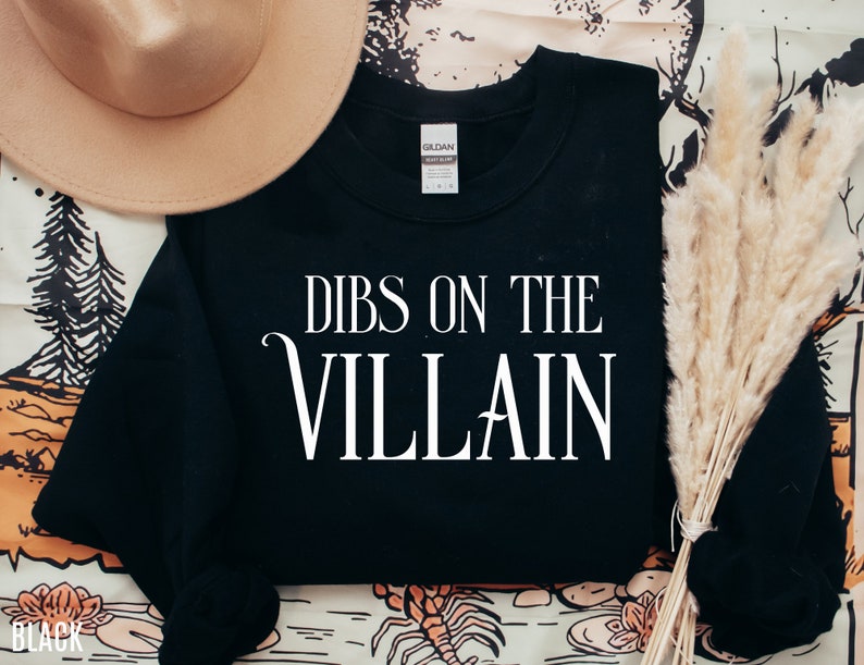 K&ouml;nnte beinhalten: Schwarzes Sweatshirt mit dem wei&szlig;en Text "DIBS ON THE VILLAIN". Das Sweatshirt ist von der Marke Gildan. Ein beiger Hut und getrocknetes Ziergras befinden sich im Hintergrund.