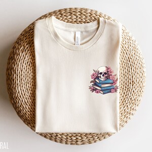 May include: Cream-colored t-shirt with a crew neck. The shirt features a graphic of a skull atop a stack of blue books, surrounded by pink flowers. The word "NATURAL" is printed at the bottom left.
