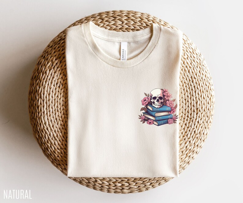 May include: Cream-colored t-shirt with a crew neck. The shirt features a graphic of a skull atop a stack of blue books, surrounded by pink flowers. The word "NATURAL" is printed at the bottom left.