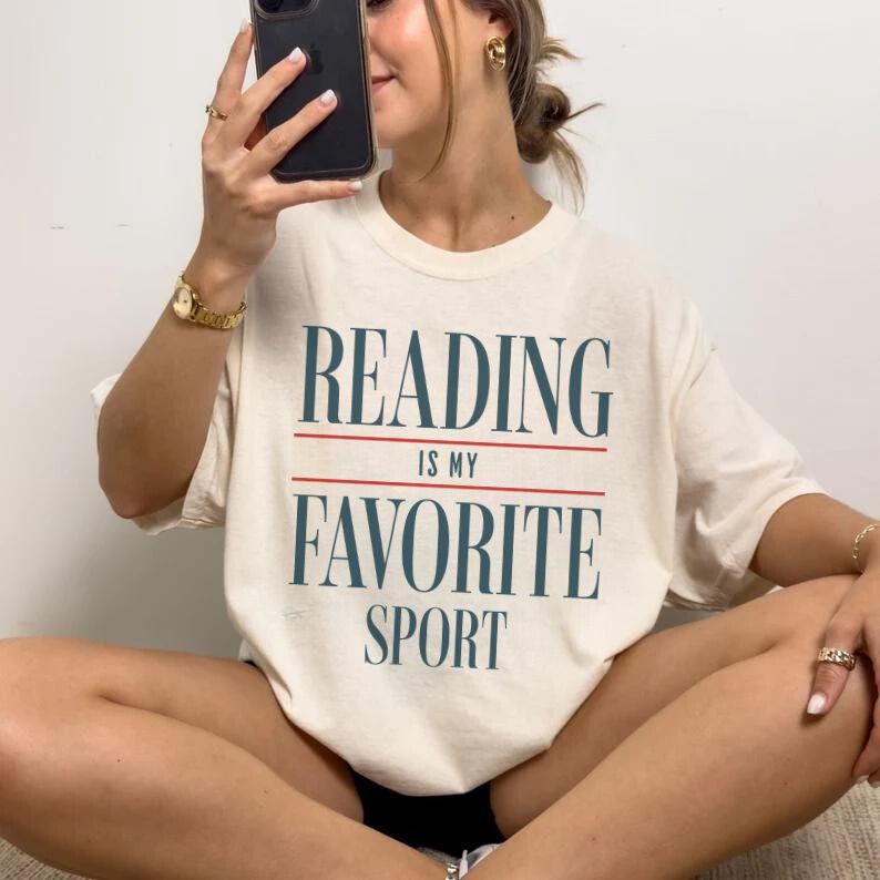 Może przedstawiać: Biała koszulka z nadrukiem "Reading is my favorite sport" w kolorze niebieskim i czerwonym.