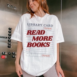 Woman wearing an oversized white Comfort Colors t shirt featuring a vintage library card graphic with bold red Read More Books lettering, photographed in an elevator for a casual reader inspired outfit and bookish lifestyle look
