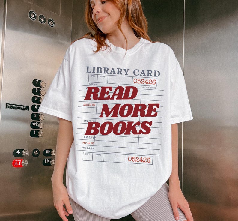 Woman wearing an oversized white Comfort Colors t shirt featuring a vintage library card graphic with bold red Read More Books lettering, photographed in an elevator for a casual reader inspired outfit and bookish lifestyle look
