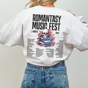 May include: Pale grey sweatshirt featuring the text "ROMANTASY MUSIC FEST 2024" and a skull design with flowers. The back of the sweatshirt lists romance novel tropes. The model is wearing blue jeans.