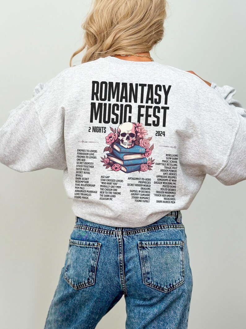 May include: Pale grey sweatshirt featuring the text "ROMANTASY MUSIC FEST 2024" and a skull design with flowers. The back of the sweatshirt lists romance novel tropes. The model is wearing blue jeans.