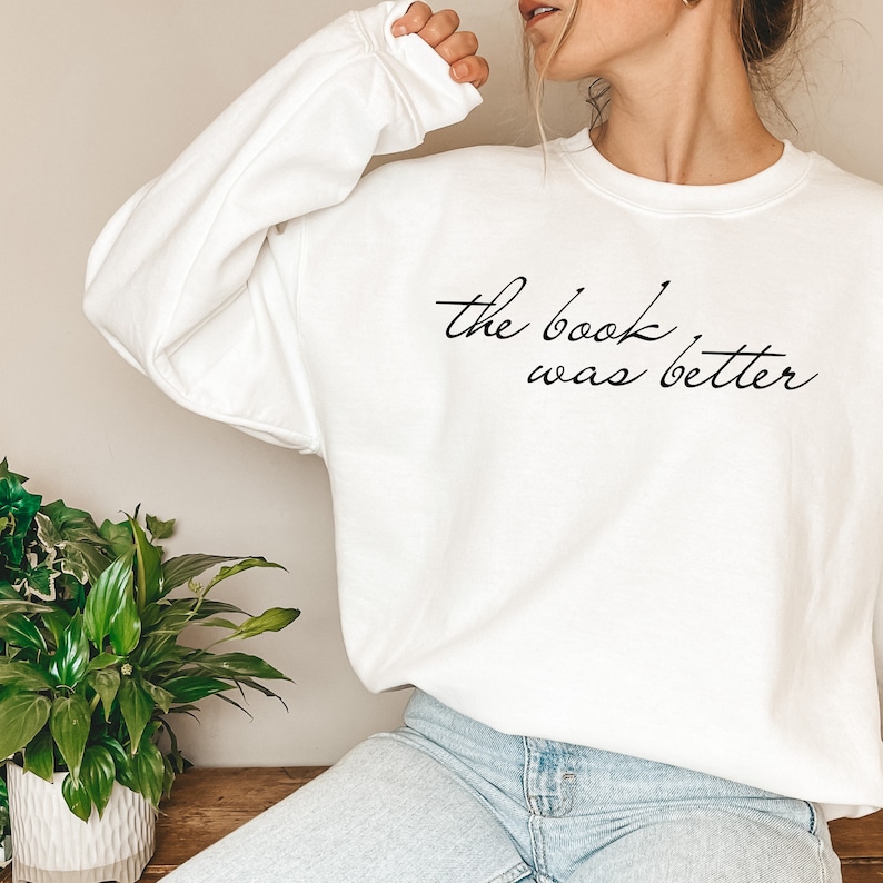 Puede incluir: Una sudadera blanca de manga larga con cuello redondo que presenta el texto en cursiva negra "the book was better" impreso en el pecho. La prenda tiene un corte holgado con pu&ntilde;os y cuello acanalados, ofreciendo un aspecto sencillo.