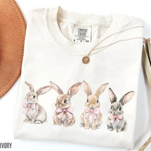 May include: Ivory-coloured t-shirt featuring four watercolour bunnies with pink bows. The shirt has a crew neck and short sleeves. A gold necklace with a pendant is draped on the shirt. The brand label "Comfort Colors" is visible.