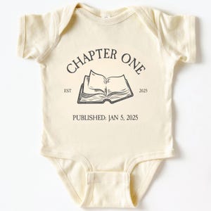 May include: A cream-coloured baby onesie featuring the text "CHAPTER ONE" above an open book illustration. Below the book, it reads "PUBLISHED: JAN 5, 2025". A newborn garment.