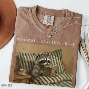 Folded espresso brown Comfort Colors t shirt with probably reading trash text and raccoon reading book artwork, styled with necklace and neutral flat lay