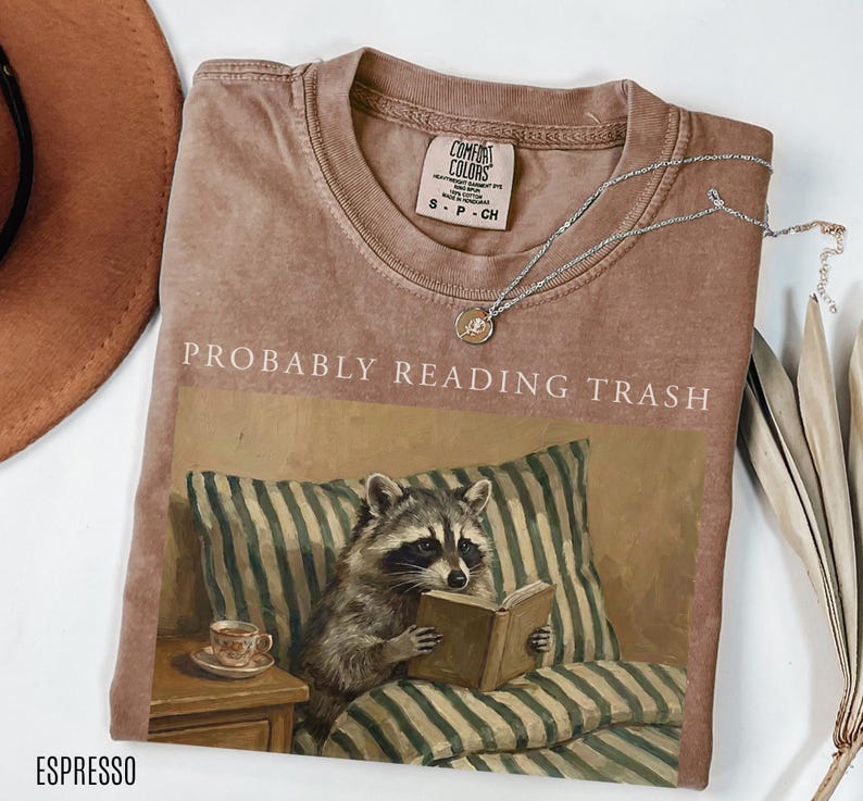 Folded espresso brown Comfort Colors t shirt with probably reading trash text and raccoon reading book artwork, styled with necklace and neutral flat lay