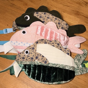 May include: Three handmade fish-shaped bags in various colors and patterns. One is dark green corduroy, another is pink with vertical stripes, and the third is a mix of floral and green velvet. Each bag has a strap with a plastic buckle.