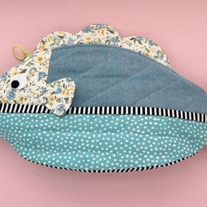 May include: A fish-shaped fabric pouch featuring a blue quilted body, a teal section with white polka dots, and floral patterned fabric on the fins and tail. It includes a black and white striped trim, a googly eye, and a bright green strap.