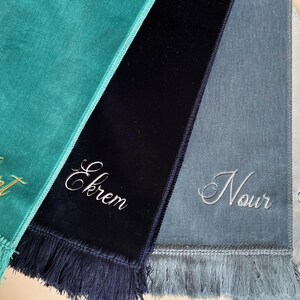 May include: Four personalised prayer rugs with fringed edges. The rugs are teal, navy blue, light blue, and light grey. The names "Albert", "Ekrem", "Nour", and "Keren" are embroidered on the rugs.