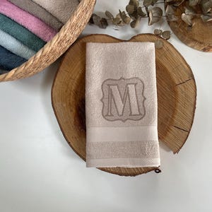 Könnte beinhalten: Ein beiges Handtuch mit einem gestickten Buchstaben "M" in einem dekorativen Rahmen. Das Handtuch ist gefaltet und auf einer Holzscheibe platziert, mit einem Korb mit gefalteten Handtüchern im Hintergrund. Das Handtuch hat einen gestreiften Rand.