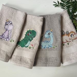 Könnte beinhalten: Vier beigefarbene Handtücher mit gestickten Dinosaurier-Motiven. Die Dinosaurier sind lila, grün, blau und braun. Jeder Dinosaurier trägt eine Krone auf dem Kopf.