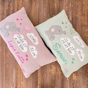 May include: Two baby pillows with embroidered details. One is pink, the other is mint green. Each features a cartoon elephant holding a heart-shaped balloon. The pink pillow has the name "LEPLA EMILIA", and the green pillow has the name "SAMUEL". Both pillows include birth details.