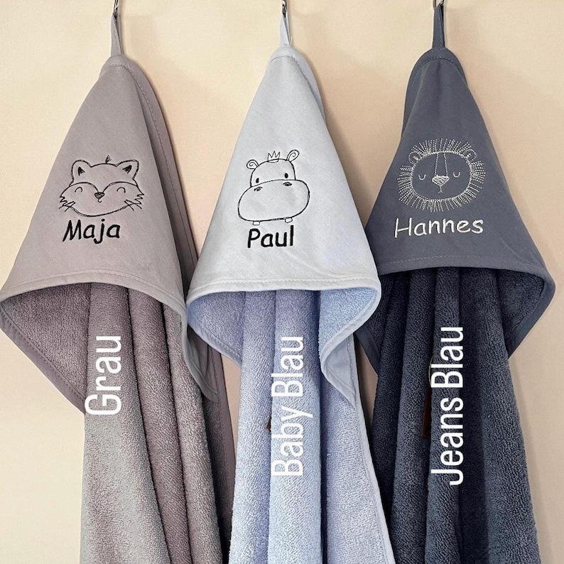 May include: Three hooded towels in gray, light blue, and dark blue with embroidered animal designs and names. The towels are labeled "Grau", "Baby Blau", and "Jeans Blau".