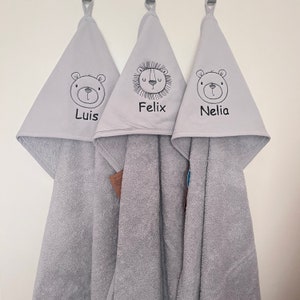 May include: Three gray hooded towels with embroidered animal faces and names. The towels feature a bear with the name "Luis", a lion with the name "Felix", and a bear with the name "Nelia".