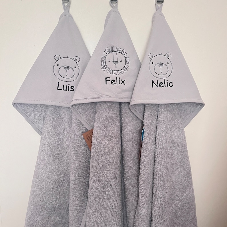 May include: Three gray hooded towels with embroidered animal faces and names. The towels feature a bear with the name "Luis", a lion with the name "Felix", and a bear with the name "Nelia".