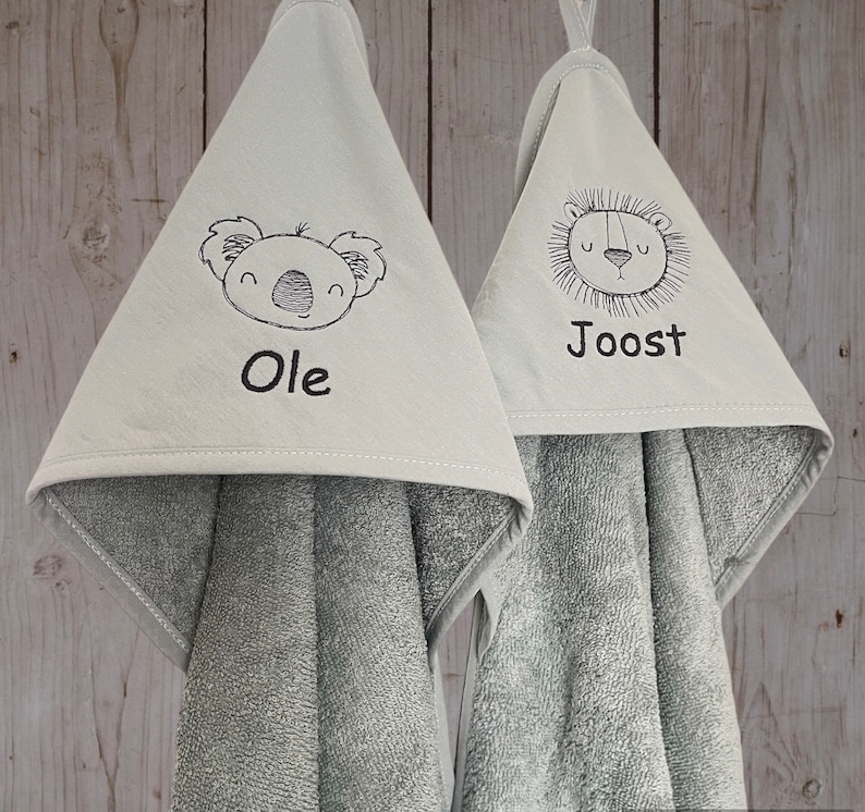 May include: Two gray hooded towels with embroidered animal faces and names. The towel on the left has a koala with the name "Ole" and the towel on the right has a lion with the name "Joost".