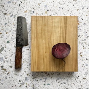 Op de afbeelding: Een houten snijplank met een rode biet erop. De plank heeft een natuurlijke houtnerf en is rechthoekig. Een mes met een zwart handvat bevindt zich links van de plank.