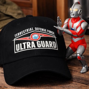 May include: Black baseball cap with the text "TERRESTRIAL DEFENSE FORCE ULTRA GUARD" in white. A red and silver action figure is in the background. The cap has a curved brim and a structured crown.