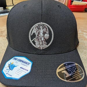 May include: Black baseball cap with a silver emblem of an angel holding a sword and shield. The cap has a curved brim and a structured crown. Includes Flexfit and Pro-Formance labels.