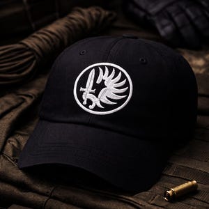 Puede incluir: Gorra de béisbol negra con un emblema blanco bordado que presenta una espada y alas. La gorra está hecha de una tela resistente y tiene una visera curva. El fondo incluye una cuerda, guantes y una vaina de bala.