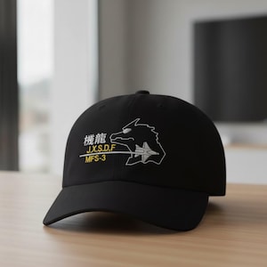 May include: Black baseball cap featuring white and yellow embroidered text and a stylized graphic. The text includes Japanese characters and the acronyms J.X.S.D.F and MFS-3. The graphic depicts a dragon-like creature and a jet.