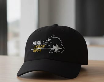 May include: Black baseball cap featuring white and yellow embroidered text and a stylized graphic. The text includes Japanese characters and the acronyms J.X.S.D.F and MFS-3. The graphic depicts a dragon-like creature and a jet.