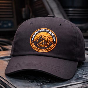 May include: Black baseball cap with a curved brim. The cap features a circular patch with the text "Jupiter Mining Corporation" and a mountain graphic in orange and white. The hat is made of a durable fabric.