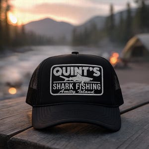 Puede incluir: Gorra de camionero negra con un parche rectangular blanco con el texto "QUINT'S SHARK FISHING Amity Island" y una ilustración de tiburón. La gorra tiene una parte trasera de malla y una visera curva. El fondo muestra una escena borrosa al aire libre.