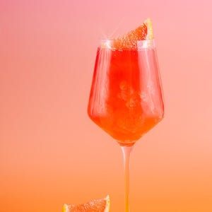 Może przedstawiać: Wysoki kieliszek wypełniony żywym pomarańczowym koktajlem, udekorowany plasterkiem grejpfruta. Drugi plasterek grejpfruta leży na powierzchni. Tło to gradient od różu do pomarańczu, tworzący ciepłą, zachęcającą estetykę.