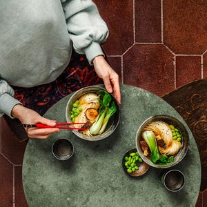 Op de afbeelding: Twee kommen ramennoedels met boerenkool, champignons en edamame op een groene tafel. Een persoon houdt eetstokjes en een kom ramen vast.