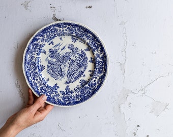 May include: Two blue and white porcelain plates with a floral pattern and a central design of a house and trees. The plates are on a white textured background.