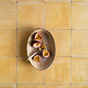 May include: A ceramic oval dish with four halved passion fruits and a gold spoon. The dish is on a yellow tiled surface.