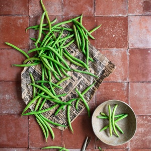 May include: A bunch of green string beans on a piece of brown and white newspaper with a bowl of green string beans on a brown surface.
