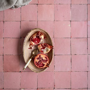 Puede incluir: Tres mitades de granada en un plato ovalado marrón claro con una cuchara de oro sobre una superficie de azulejos rosa.