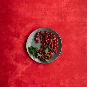 Könnte beinhalten: Ein Metallteller mit leuchtend roten Beeren und grünen Blättern auf einem leuchtend roten Hintergrund. Die Beeren sind gruppiert, und die Blätter sorgen für einen frischen Kontrast. Die Aufnahme ist von oben.