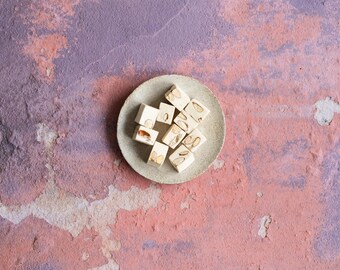 May include: A small white ceramic plate with a pile of nougat candy. The nougat is white and has a light brown center. The plate is on a textured pink and purple background.