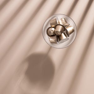 Könnte beinhalten: Draufsicht auf einen klaren Glasteller mit mehreren goldfarbenen Kaffeekapseln. Die Kapseln sind in einem Haufen angeordnet. Der Hintergrund ist eine hellbraune Oberfläche mit Schatten.