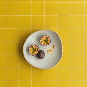 May include: A white ceramic plate with two halves of a passion fruit and a whole passion fruit. The plate sits on a yellow tiled surface. The passion fruit halves reveal yellow pulp and black seeds.