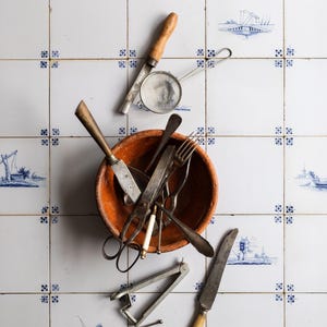 May include: A collection of vintage kitchen utensils, including a wooden handled strainer, a pair of scissors, a metal nutcracker, a wooden handled knife, and several forks and spoons, are arranged in a terracotta bowl on a white tiled surface with a blue and white pattern.