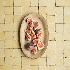 Może przedstawiać: Owalny, nakrapiany beżowy półmisek ceramiczny z kilkoma pokrojonymi świeżymi figami. Figi mają ciemnofioletową skórkę oraz żywe, różowe wnętrze z nasionami. Półmisek znajduje się na środku siatki złożonej z jasnych, żółtych płytek.