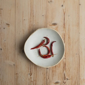 Puede incluir: Un plato de cerámica blanco, de forma irregular, con chiles rojos dispuestos sobre una superficie de madera clara. Los chiles están colocados para parecer las letras "BI". El plato tiene una textura moteada.