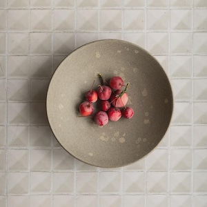 May include: A round, speckled, gray ceramic plate holds a cluster of small, red crabapples with green stems. The plate sits on a light-colored, square-tiled surface, creating a minimalist, natural aesthetic.