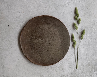 May include: A round, brown ceramic plate with a textured surface. The plate is set against a grey background, with a sprig of green grass to the right. The plate is approximately 25 cm in diameter.