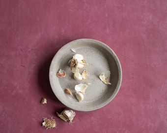 May include: A small, gray ceramic bowl filled with peeled garlic cloves. The bowl is sitting on a dark red surface.