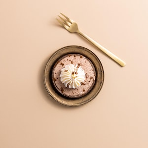 Könnte beinhalten: Ein kleines Schokoladendessert mit Schlagsahne auf einem goldfarbenen Teller, neben einer goldenen Gabel. Das Dessert befindet sich auf einer hellbraunen Oberfläche.